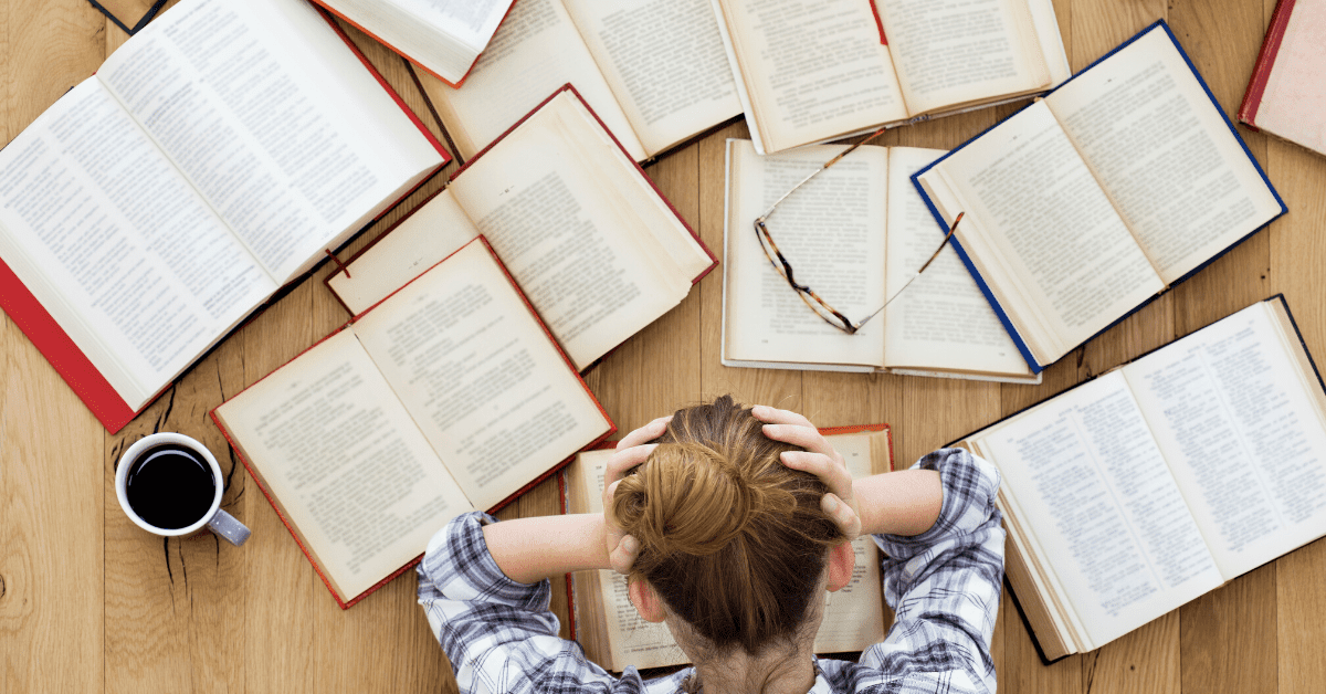 jeune femme dépassée par la charge d'étude qui se prend la tête dans les mains devant une table remplie de livres